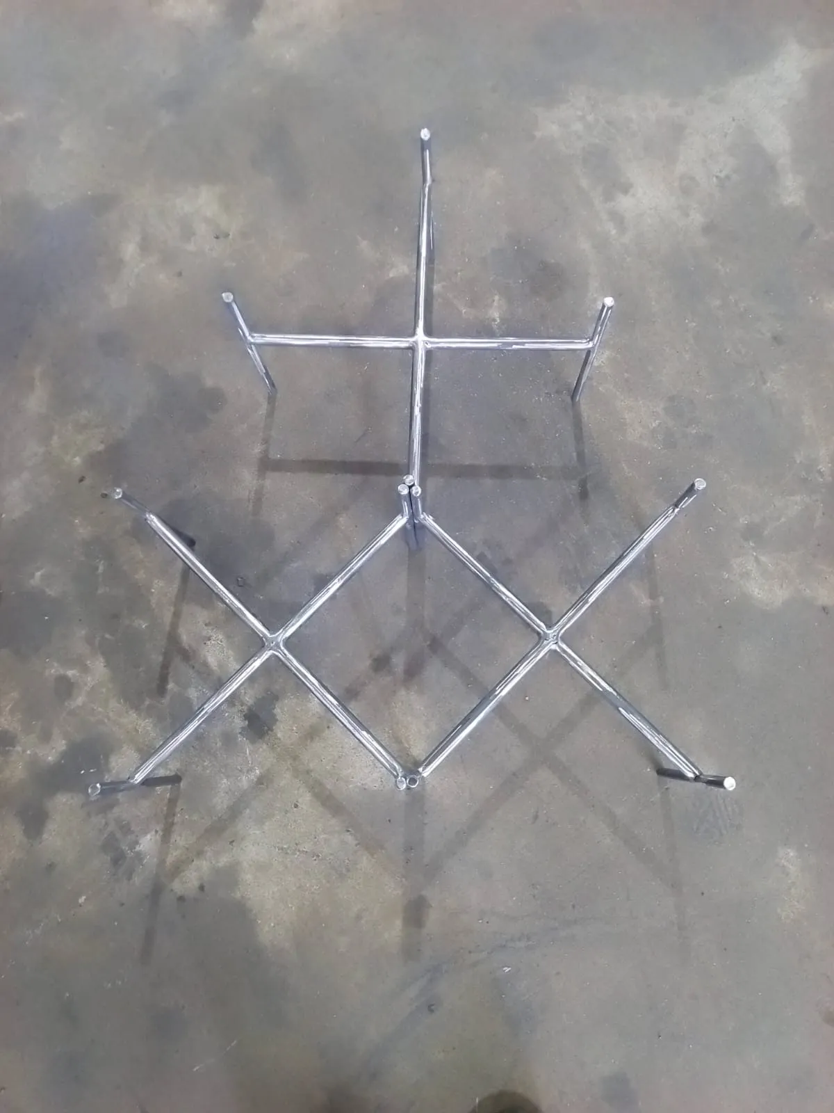 Cross-braced stainless steel table base laid flat on the workshop floor, showing the welded centre joint